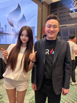 A young woman with long brown hair in a cream sweater stands next to a smiling man in a dark suit, both making finger hearts in front of a cityscape poster.