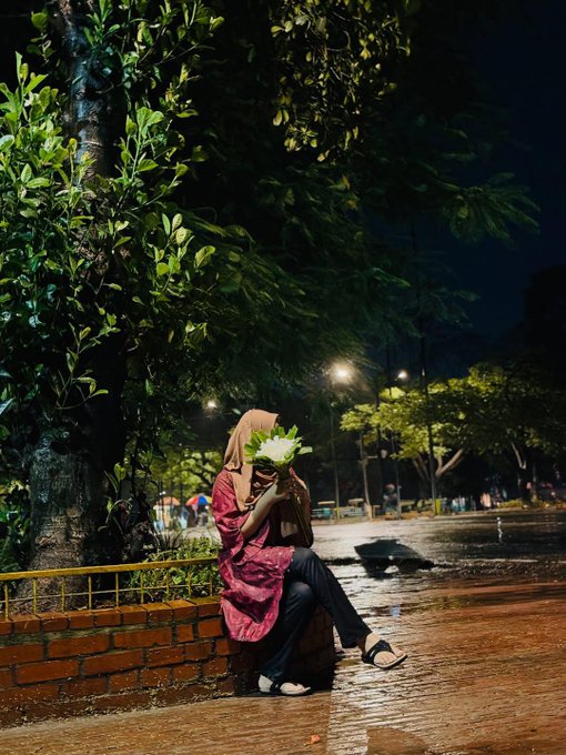 A nighttime photo of a person in a hijab and pink garment sitting on a brick curb holding a bouquet of flowers that obscures their face, on a wet street under streetlights.