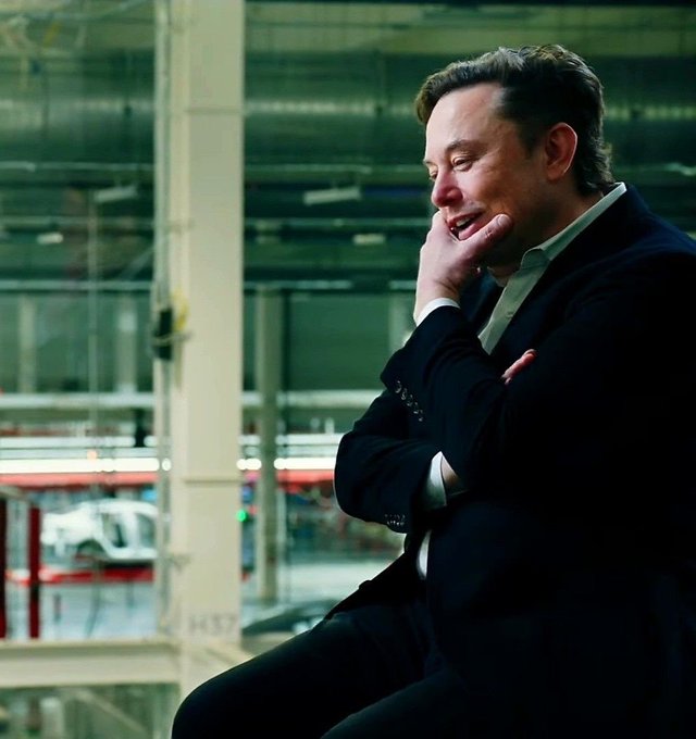 Elon Musk in a dark suit laughing with his hand on his chin in what appears to be a Tesla factory setting with vehicles visible in the background.