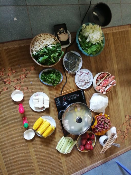 A top-down view of a hot pot meal spread on a bamboo mat, featuring a central pot surrounded by various bowls of fresh vegetables, bean sprouts, tofu, corn, meat slices, and dipping condiments.