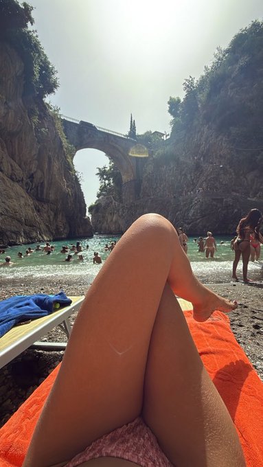 A first-person view of someone relaxing on an orange towel at a beach, showing their crossed legs with swimmers and a stone bridge in the background.