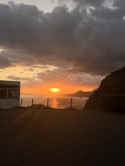 A sunset scene over water viewed from a paved overlook, with dramatic cloudy skies, a setting sun, and a coastal landscape visible in the distance.
