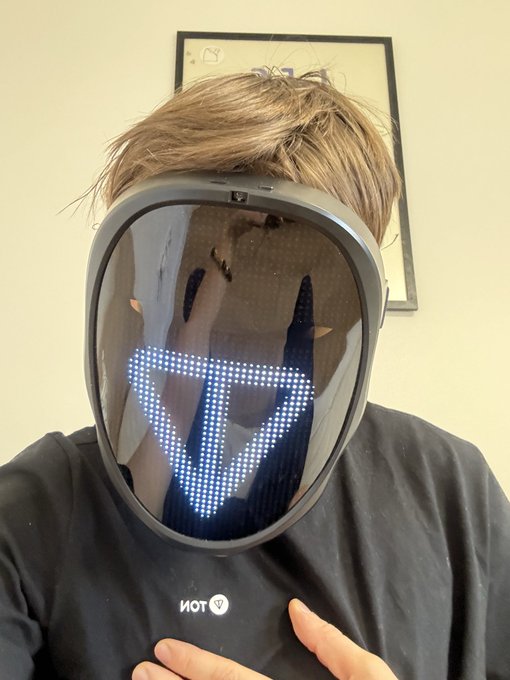 The image features a person wearing a mask with a triangle symbol, possibly representing a robot or futuristic character. They are posing for a photo while holding their hand up to the camera. The caption of the image reads "GM to the TON believers". This suggests that the person is likely a fan of GM (General Motors) and might be interested in futuristic technology or robotics.
