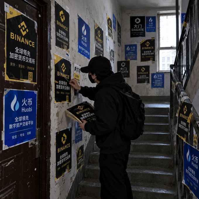 A person wearing a black cap, face mask, and backpack stands on a stairwell covered with cryptocurrency exchange posters for Binance, Huobi, and DAGATE.