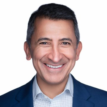 A professional headshot of a smiling man with dark hair, wearing a navy blazer over a checkered shirt, against a white background—appearing to be Faryar Shirzad, Coinbase's chief policy officer, as referenced in the accompanying post about bipartisan support for the CLARITY Act.