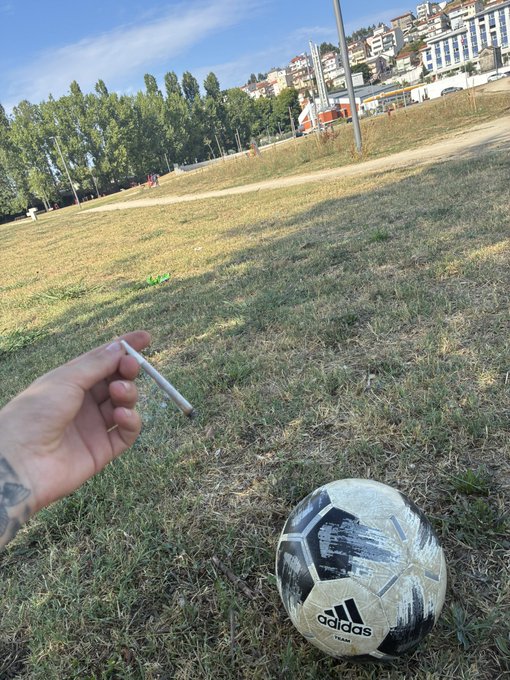 A tattooed hand holding a cigarette is shown over a grassy field, with a black and white Adidas soccer ball resting on the ground.