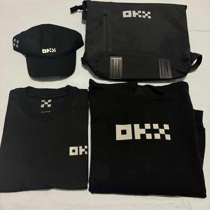 A merchandise collection featuring a black crossbody bag, black cap, black t-shirt, and black sweatshirt, all branded with the 'OKX' logo in white.
