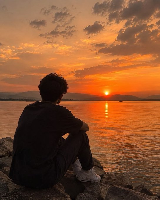 A silhouetted figure sits on rocky shores gazing out at a golden sunset reflecting across calm waters toward distant mountains. The tranquil scene of quiet reflection mirrors the contemplative, end-of-day tone of the post about Nikita Bier's resignation and a return home.