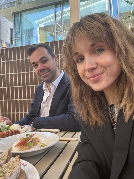 A selfie of a woman with wavy brown hair in a black blazer smiling at an outdoor restaurant table, with a man in a dark suit seated behind her next to plates of food including pasta.