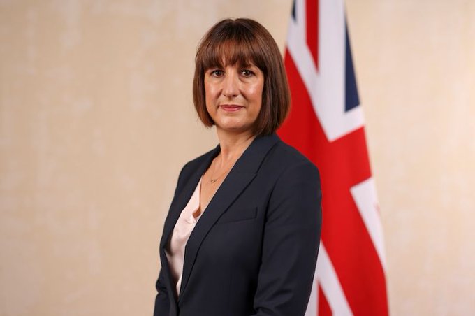 A woman with shoulder-length brown hair wearing a dark blazer over a white top, standing in front of a Union Jack flag.