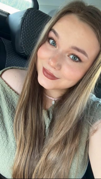 A young woman with long blonde hair and blue eyes smiles softly in a car selfie, wearing a sage green off-shoulder top and a pink choker necklace. The image radiates hopeful optimism, reflecting her heartfelt dream of one day seeing all her artworks on Transient completely sold out.