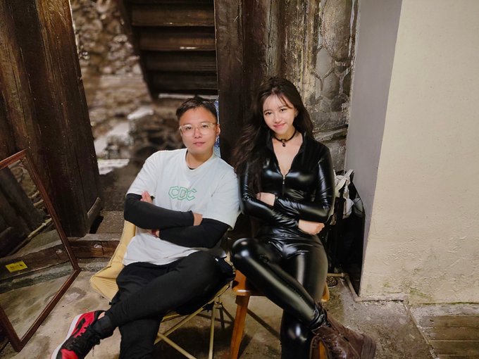 Two people sit side by side in a rustic wooden interior; the man wears a white "CDC" t-shirt and glasses, while the woman beside him is dressed in a black leather outfit with a choker. The image accompanies a post about visiting Dali to meet the "K-line Guru" @Paris13Jeanne, where the man appears to be learning trading strategies and experiencing @protocol_fx @FX100Perp's testnet product, with the woman—presumably the trading guru—sitting beside him in a posed yet stylish setting that reflects the post's mix of humor and finance talk.