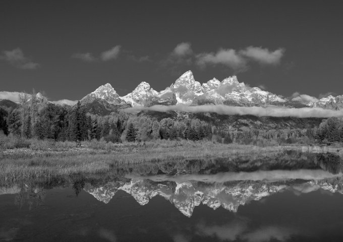 A black and white landscape photograph of snow-capped mountains reflected in a calm body of water, framed by trees and clouds.