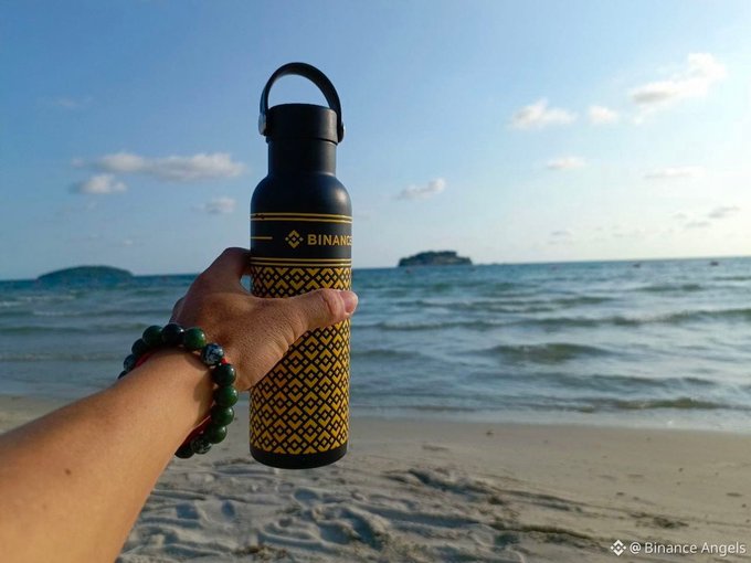 A hand wearing a beaded bracelet holds up a black and gold 'BINANCE' branded water bottle on a beach with the ocean and sky in the background.
