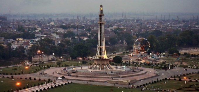 An aerial view of the Minar-e-Pakistan monument in Lahore at dusk, with a Ferris wheel and cityscape visible in the background.