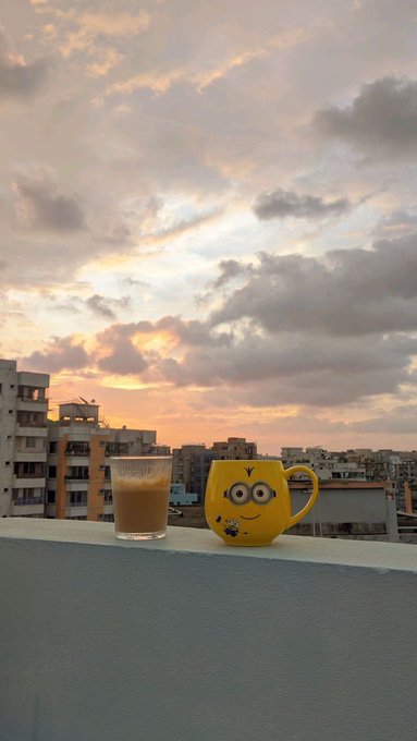 A serene sunset scene on a balcony or rooftop features a glass of milk or coffee beside a yellow mug decorated with a smiley face character, overlooking a city skyline under dramatic pink and gray clouds.