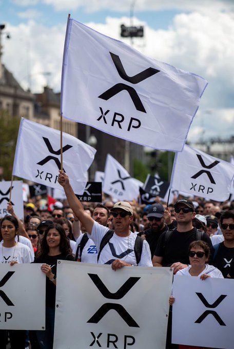 The image shows a large group of people holding up white flags with black text that reads "XRP". There are at least six visible signs, each displaying the word "XRP" prominently. The crowd appears to be gathered for an event or demonstration related to cryptocurrency and XRP specifically.