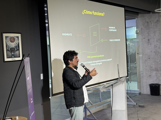 A man in a dark jacket speaks into a microphone while presenting a slide titled '¿Cómo funciona?' which diagrams the relationship between Hasrate, Hashrate Comparator, and a NWC payment coordinator.