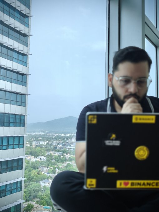 A bearded man with glasses working intently on a laptop covered in cryptocurrency and Binance stickers, sitting by a large window overlooking a cityscape with mountains in the distance.