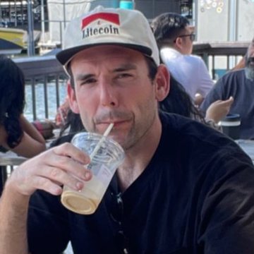 A man wearing a "Litecoin" baseball cap drinks an iced coffee beverage through a straw at an outdoor cafe.