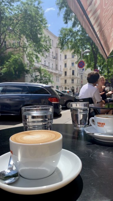 A frothy cappuccino and two water glasses sit on a café table with a view of a sunny European street, capturing the start of a leisurely morning. The saucer reads "POLLER KAFFEE," suggesting a stop at a Viennese-style coffeehouse to enjoy the day.