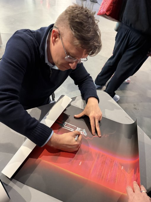 A man wearing glasses and a dark sweater signing posters or artwork with a marker at what appears to be a signing event or convention.