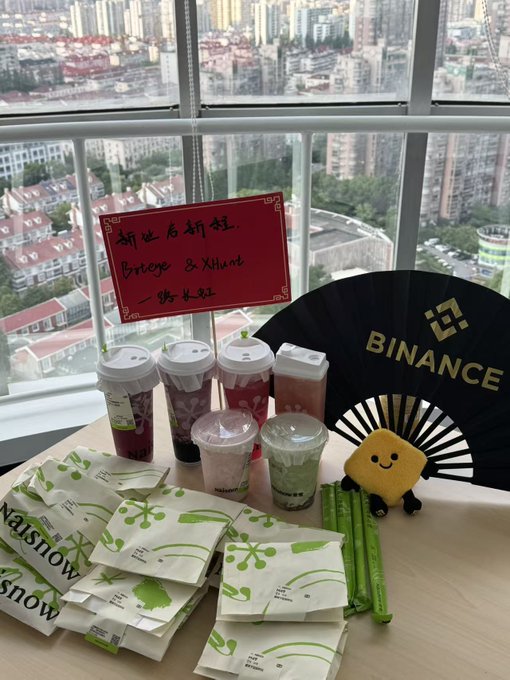 A welcome snack arrangement on a high-rise table featuring Mixue branded items including green tea and other drinks in cups, packaged snacks, a Binance folding fan, and a card with Chinese text welcoming 'Brieye & Xihuan'.