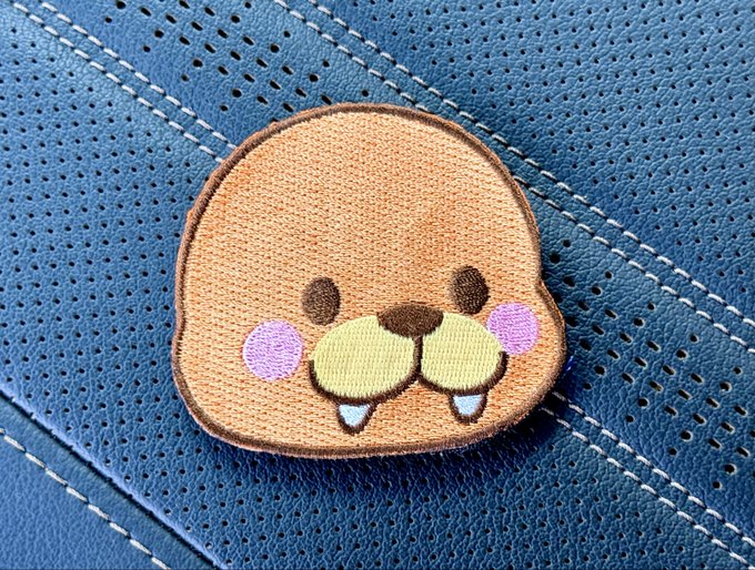 A small embroidered patch of a cute cartoon seal or sea lion face with pink cheeks, two small teeth, and a green nose resting on blue perforated leather material.