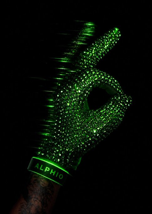 A hand wearing a glowing green glove makes a peace sign in the dark; the glove reads 'ALPHIO' on the wristband and appears to be made of many sparkling green jewels.