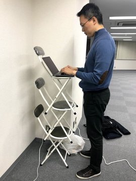 A man in a blue sweater and glasses stands at a folding chair setup, working on a laptop placed on the chair in what appears to be an office or conference space.
