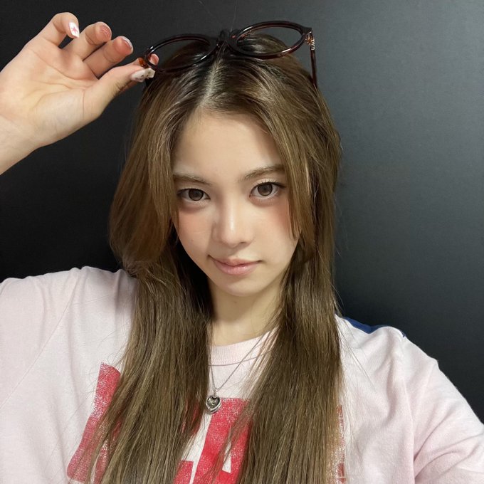 A young woman with long light brown hair and a pink sweatshirt holds sunglasses on top of her head while looking directly at the camera.