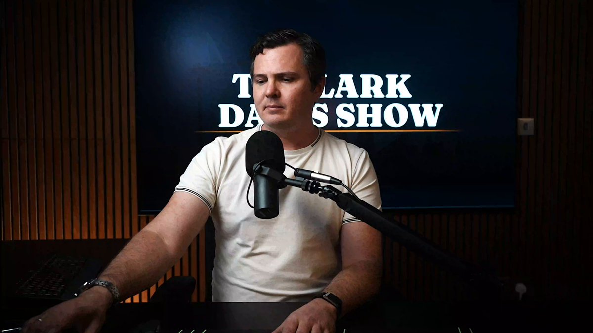 The image features a man wearing a white shirt, sitting at a desk with a microphone in front of him. He appears to be speaking into the microphone or recording a podcast. The background shows a TV screen displaying "The Clark D. Show" along with some text and logos related to cryptocurrency and stock market updates.