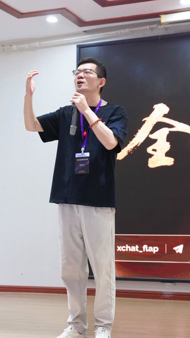 A young man wearing glasses and a black t-shirt holding a microphone and gesturing while speaking at what appears to be an xchat_fiap event.