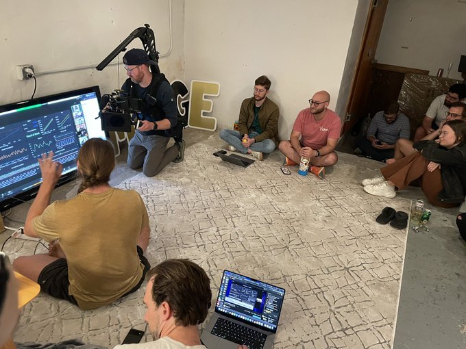 A group of filmmakers and crew sit cross-legged on a stone-patterned floor while monitoring footage on a large screen and laptop, suggesting a behind-the-scenes moment from a video production. A large decorative "GE" sign sits against the wall, hinting at the project's branding or name being created four years ago today.