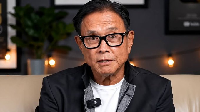 Close-up of an older man with black-framed glasses wearing a dark blazer over a white t-shirt, speaking in what appears to be a video interview setting.