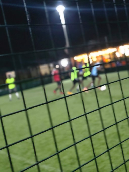 A blurry nighttime photo taken through a chain-link fence, showing people playing soccer on a lit turf field.