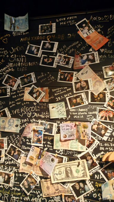 A dark wall covered with handwritten messages, signatures, polaroid photos, and various currencies pinned up as memorabilia.
