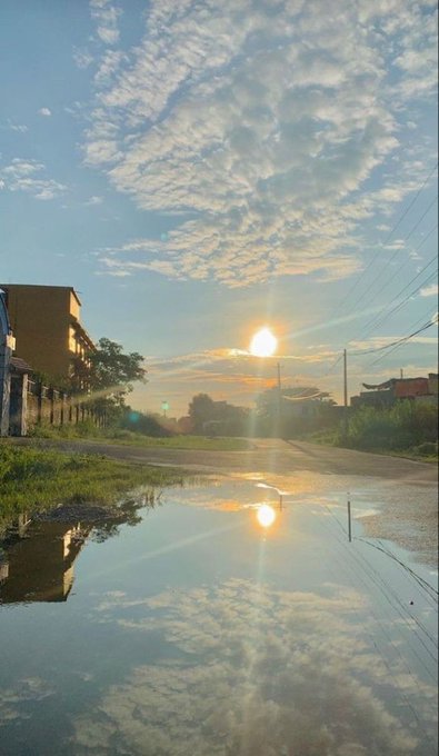 Scenic sunset photo over a wet rural road with the sun's reflection in a puddle, surrounded by clouds and silhouettes of houses and utility poles.