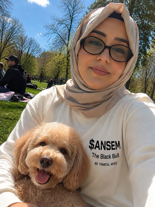 This image is of a girl smiling and posing for a selfie with her dog while wearing a long sleeved shirt that says "$ANS̩̩𝓐𝓶 The Black Bull NOT FINANCIAL ADVICE". Given that a girl is wearing a shirt that references $ANSEM, and her dog, there is a correlation to the post regarding supporting and buying a shirt for $ANSEM.