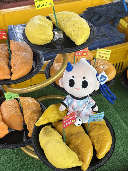 A display of durian pastries in various colors (yellow, orange, beige) arranged on black tiered trays, with a plush mascot wearing a panda-patterned outfit positioned among them. Small flags with Chinese and English text label the different varieties.