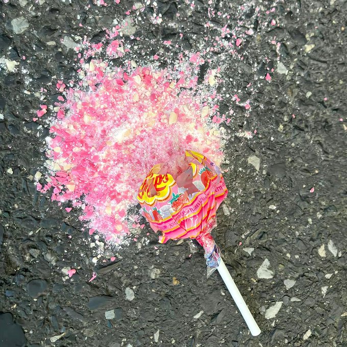 A smashed strawberry lollipop lying on wet asphalt with pink candy pieces scattered across the dark textured surface.