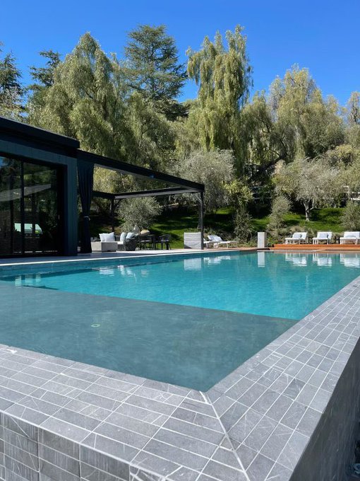 A luxurious modern backyard features an infinity-edge pool with dark tile surrounding, overlooking lush green hills under bright sunshine. The post's caption humorously suggests this upscale property is a sign to invest in real estate.