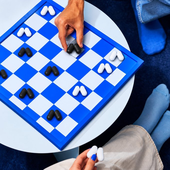An overhead view shows two people playing a creative chess game on a blue and white checkered board, using pill-shaped objects as game pieces—white pills for one player and black pills for the other.