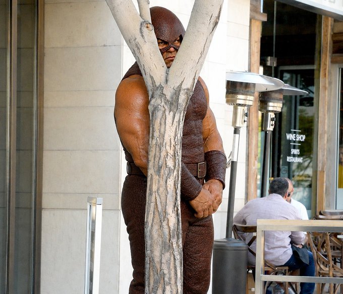 A life-sized statue of a masked superhero figure in brown leather is standing between tree branches outside a wine shop where a man sits at a table.