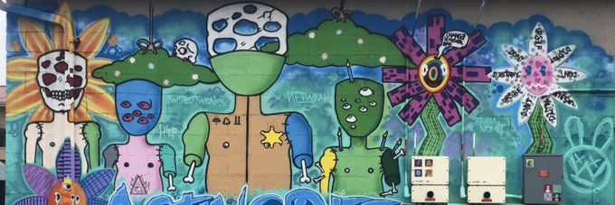 A colorful graffiti mural featuring various whimsical characters including skull-faced flowers, blue and green creatures, mushrooms, and abstract designs painted on a wall with utility boxes in front.