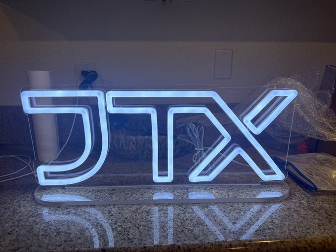 An illuminated white neon sign displaying the stylized 'JTX' logo sits on a speckled granite countertop in a dimly lit room.