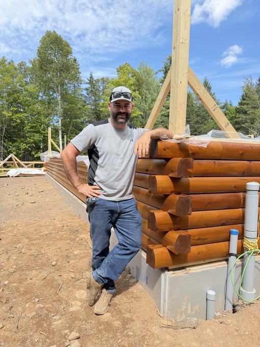 A man wearing a baseball cap and sunglasses posing proudly next to a partially constructed log cabin with wooden framing visible in the background.