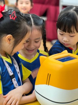 Children gathered around a yellow and white robot in what looks like a classroom setting, smiling and engaged with the device.
