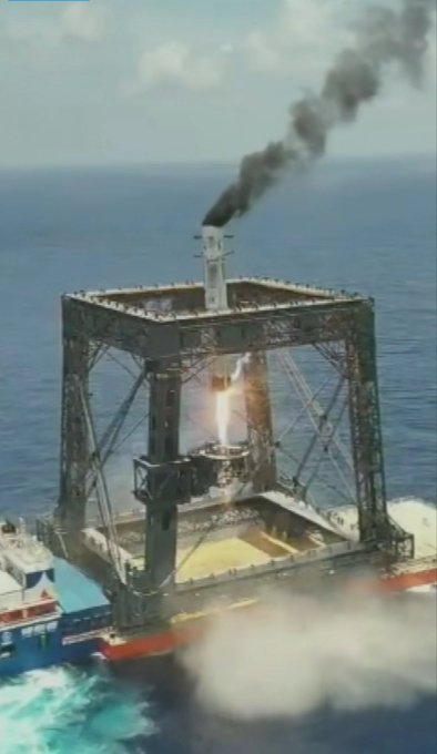 A rocket launches from a sea-based launch platform, with bright flames and dark smoke trailing from its engine as it ascends above the ocean.