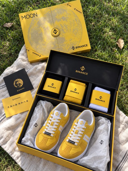 A Binance-branded yellow sneaker gift box set displayed on a picnic blanket, featuring "MOON" and "BINANCE" text along with Chinese characters "月满山海秋知人意" on the accompanying card, unboxing the festive Mid-Autumn Festival surprise the post describes receiving from Binance.